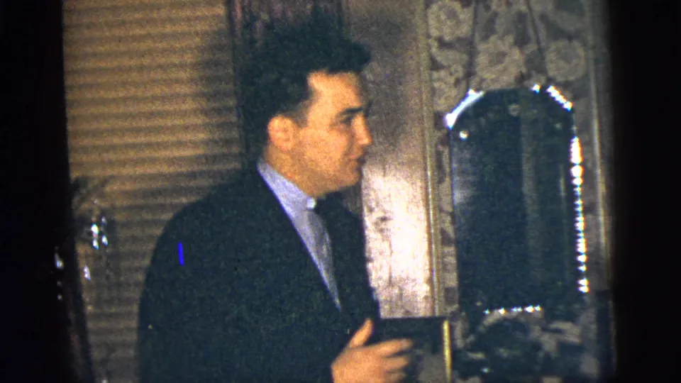 A suited adult man gestures while speaking or reacting in a 1956 New York City private interior.