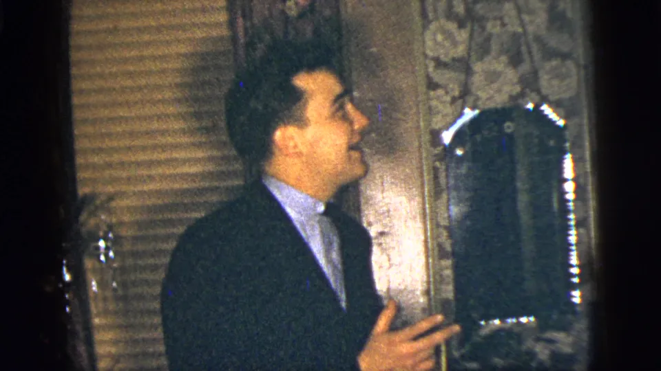 The man looks upward and gestures in the dim interior, with blinds and patterned wallpaper visible behind him.