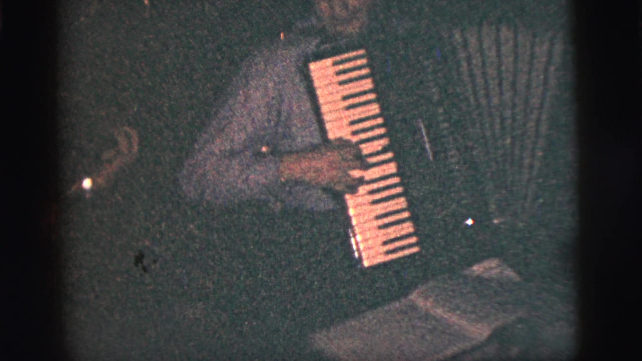 In 1956 New York City home-movie footage, an adult man plays a button accordion in a dark interior with visible grain and soft focus.