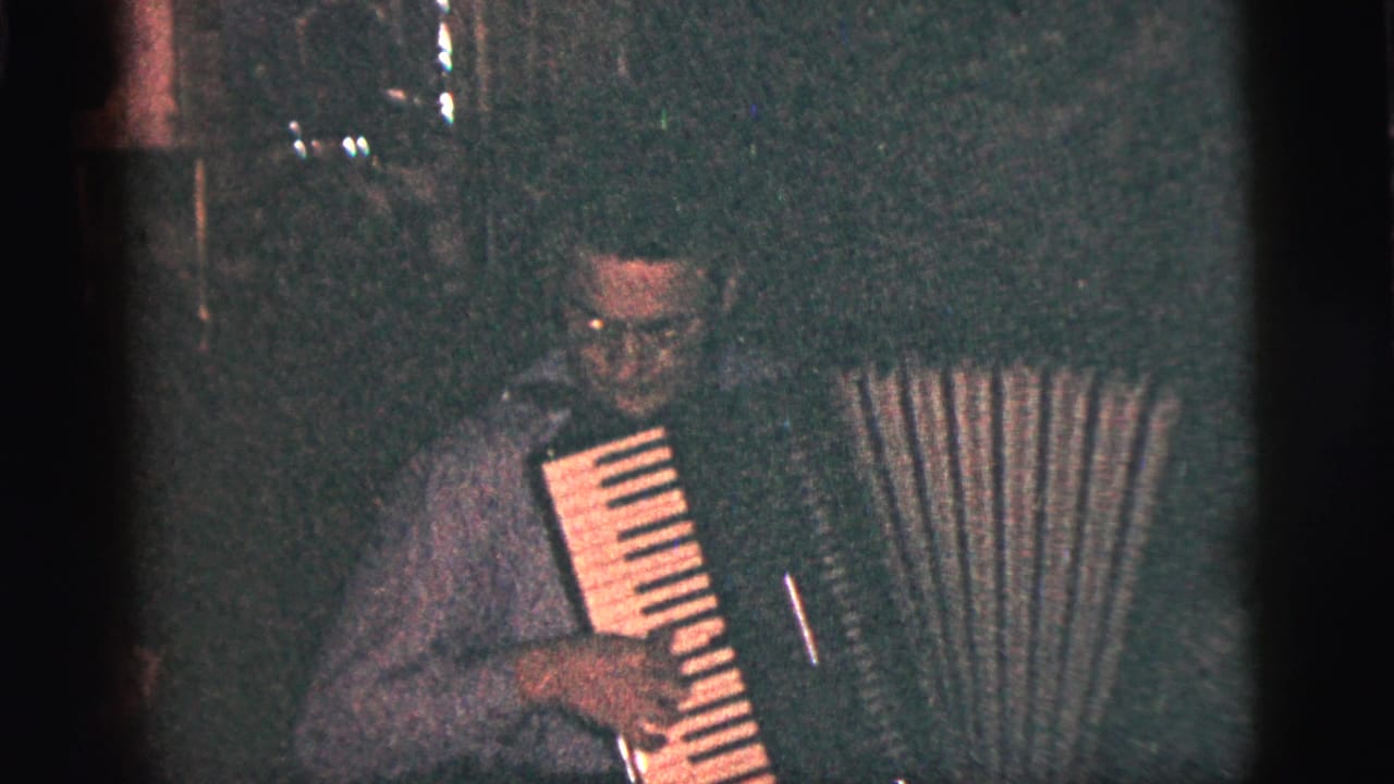 The accordion player looks downward toward the keys and bellows in a low-light 1956 New York City indoor home-movie frame.