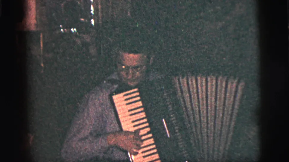 The accordion player looks downward toward the keys and bellows in a low-light 1956 New York City indoor home-movie frame.