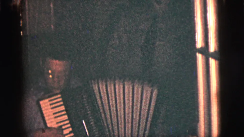 A grainy 8mm frame shows the man and accordion partly cropped, with bellows and a bright vertical film edge visible.