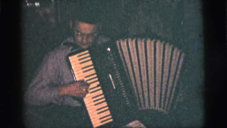 In supplied 1956 New York City 8mm footage, a man looks down while playing an accordion in a dim interior.