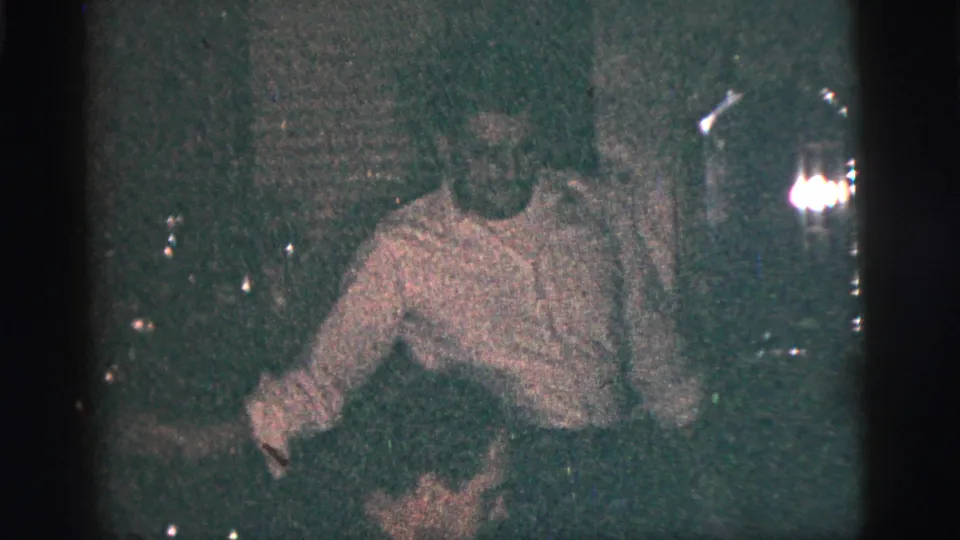 A person in a light shirt appears seated or leaning in the same dim indoor setting, with other figures partly visible nearby.