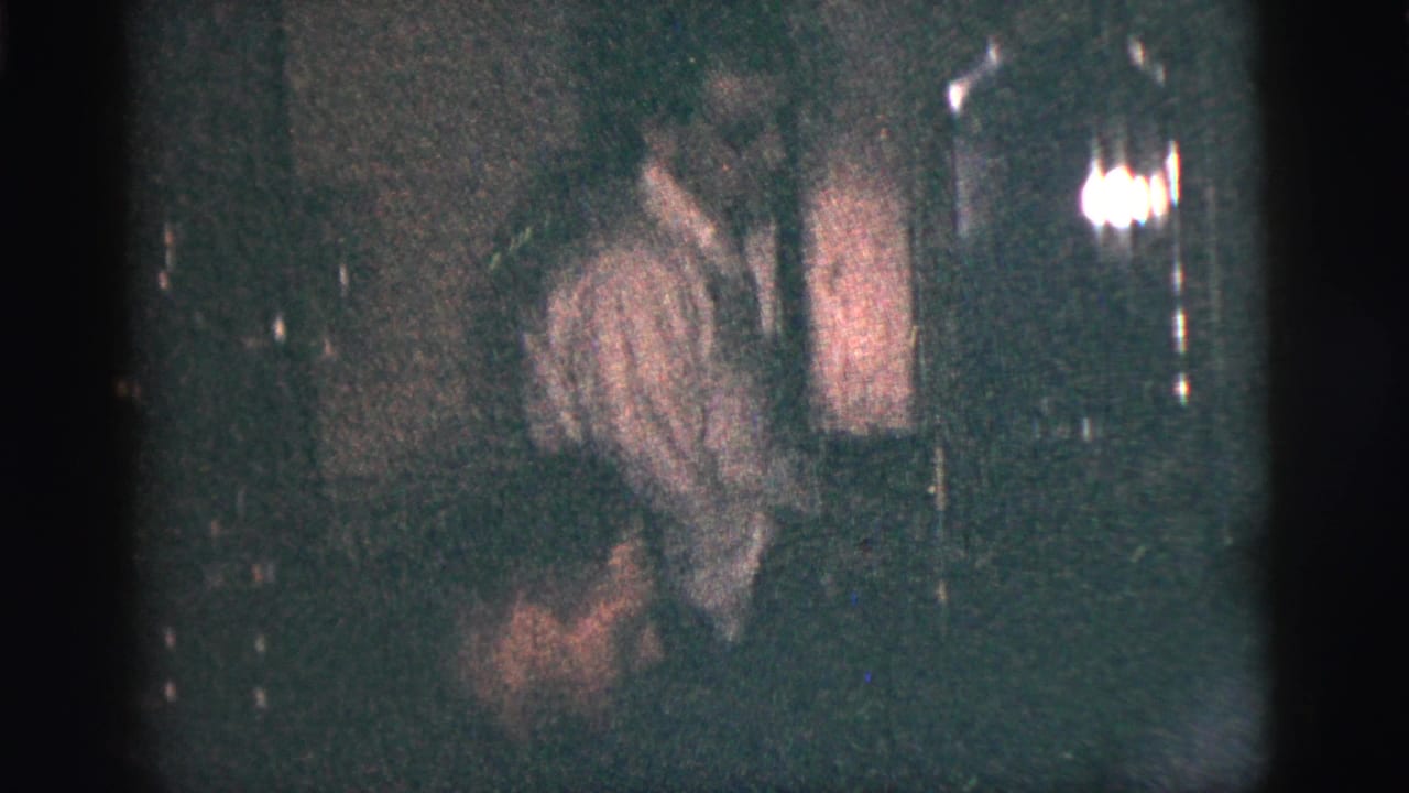 Young men sit in a dim interior, one in a light shirt and tie partly facing the camera.