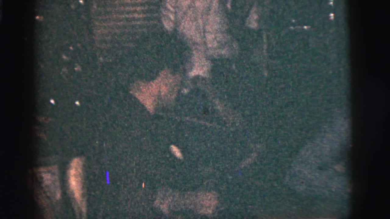 Grainy 1956 New York City interior frame showing a man leaning forward among seated friends in low light.