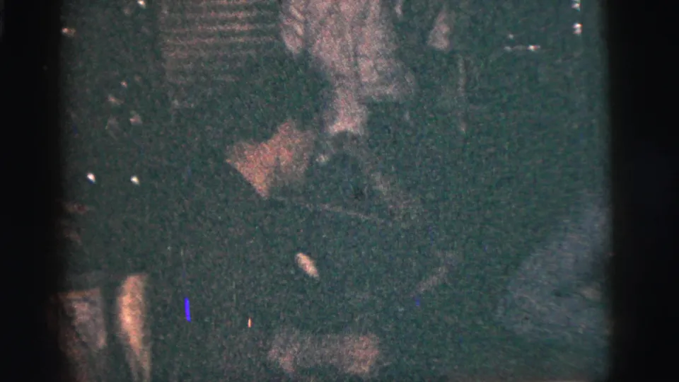 Grainy 1956 New York City interior frame showing a man leaning forward among seated friends in low light.