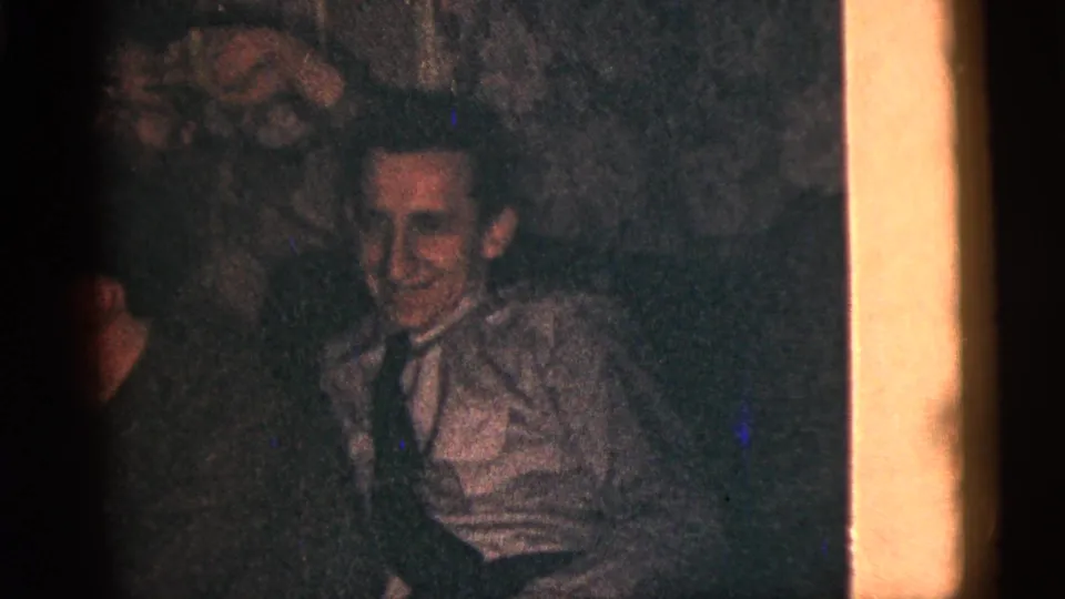 1956 New York City home-movie still of a smiling young man in a light shirt and tie looking toward the camera among friends indoors.
