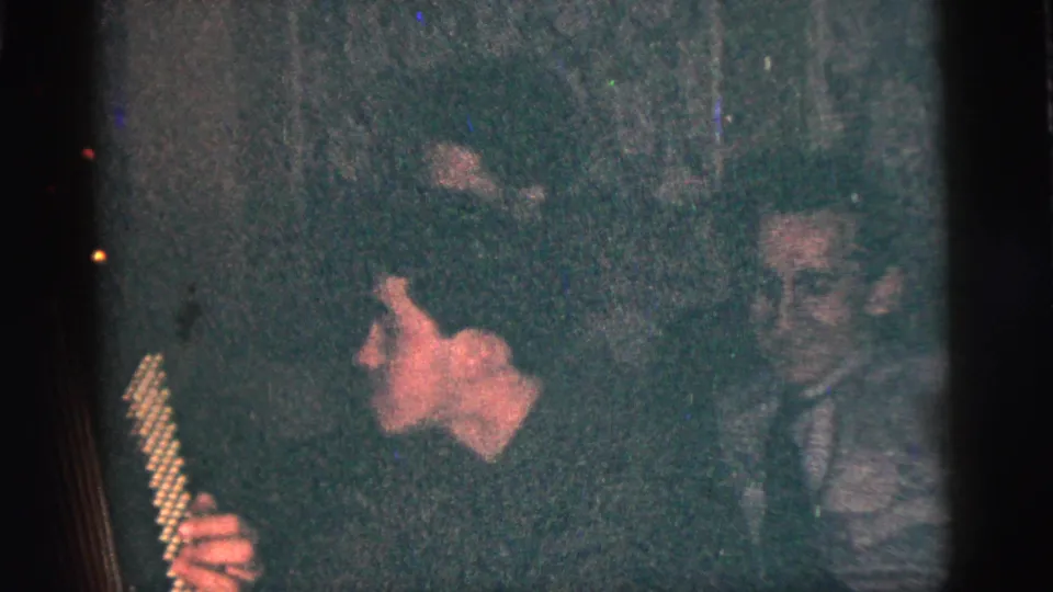 1956 New York City frame showing a young man holding a small stringed instrument as another man sits nearby in a dark private interior.