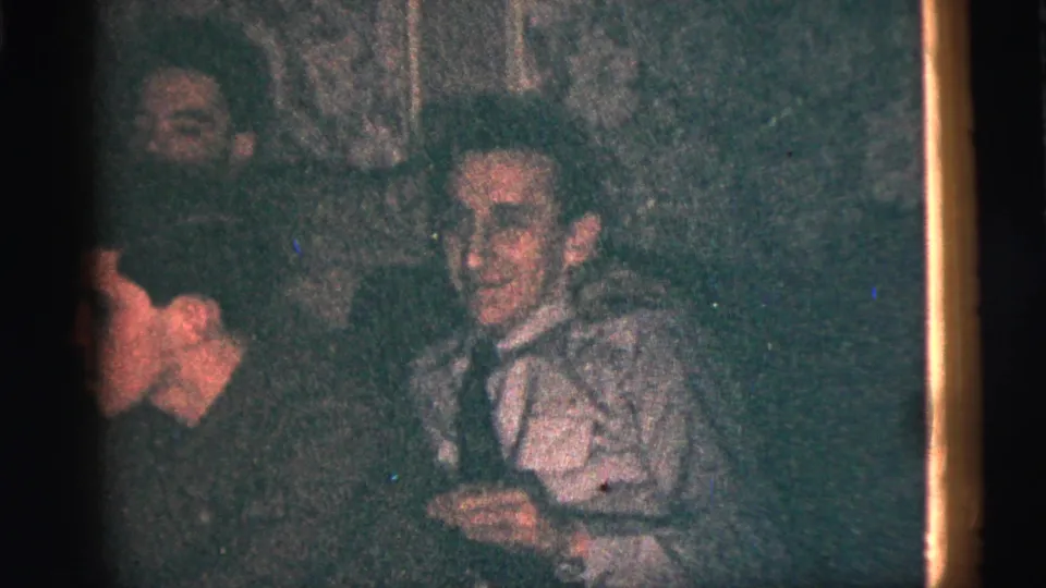 1956 New York City home-movie frame showing a smiling adult man near another guest during an indoor gathering.