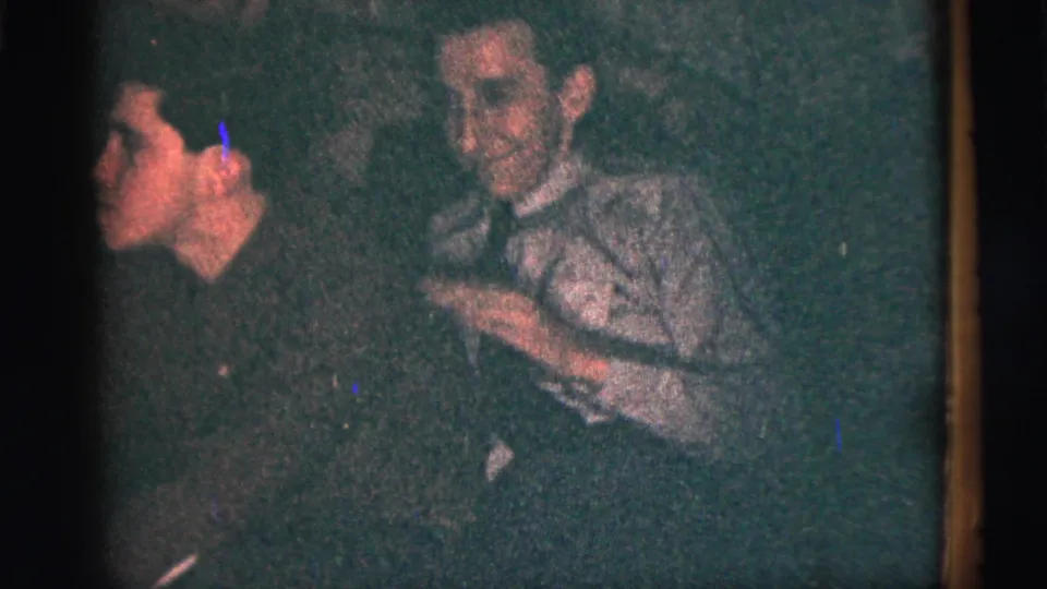 1956 New York City home-movie frame of two adult men in close conversation or reaction during a low-light party scene.