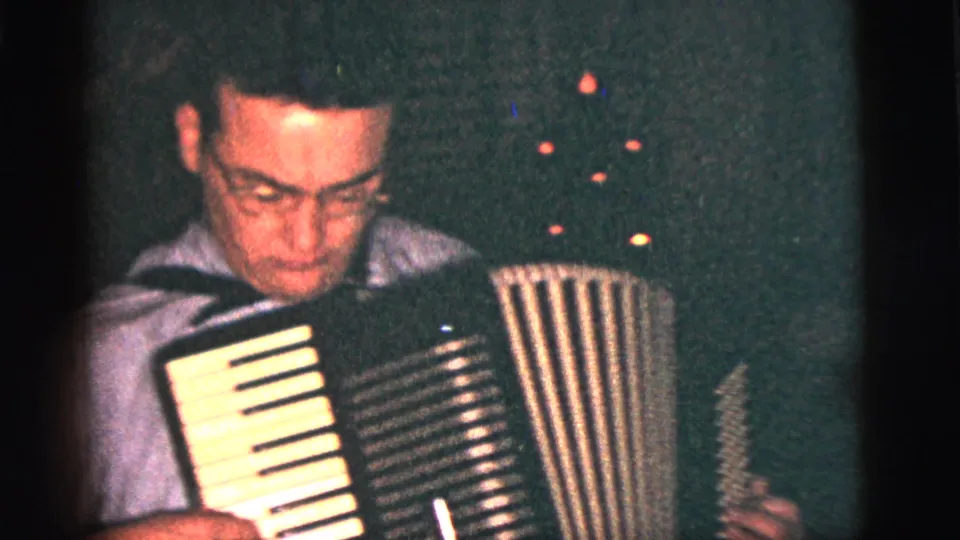 1956 New York City home-movie frame showing an adult man focused on an accordion in a shadowed private interior.