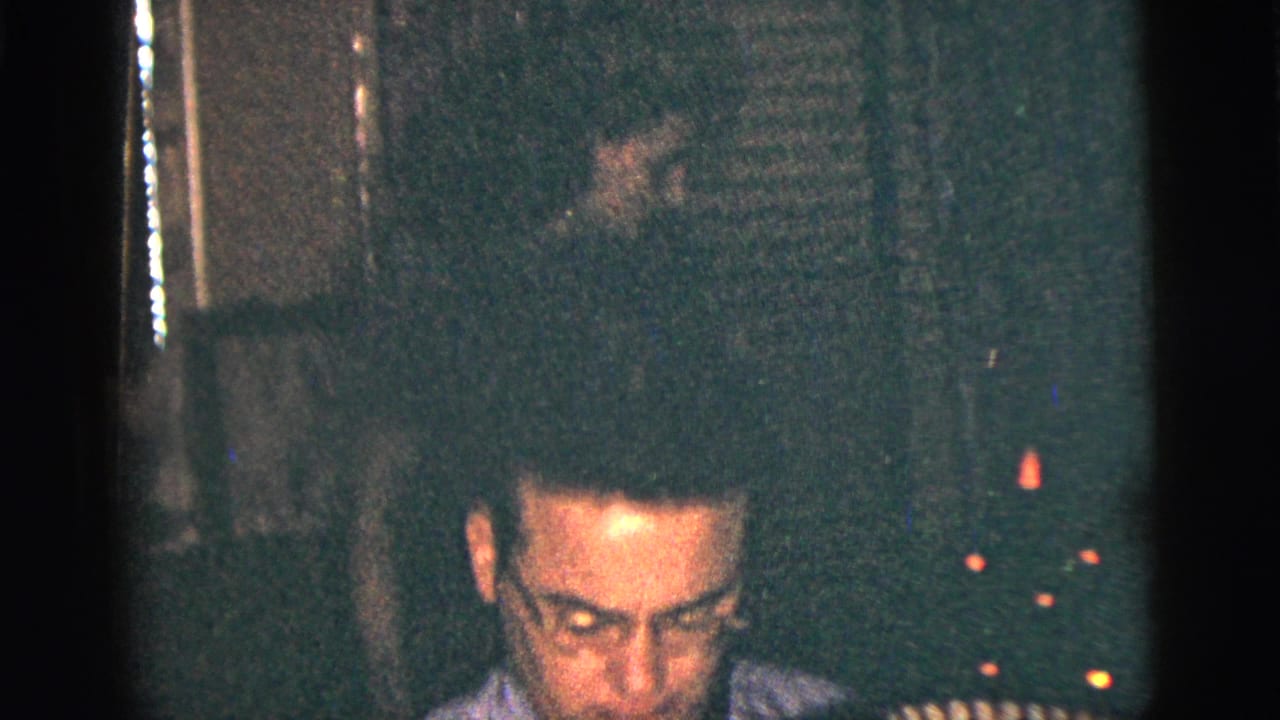 The camera catches the accordion player low in frame, with shadowed background detail and warm points of light in the private room.