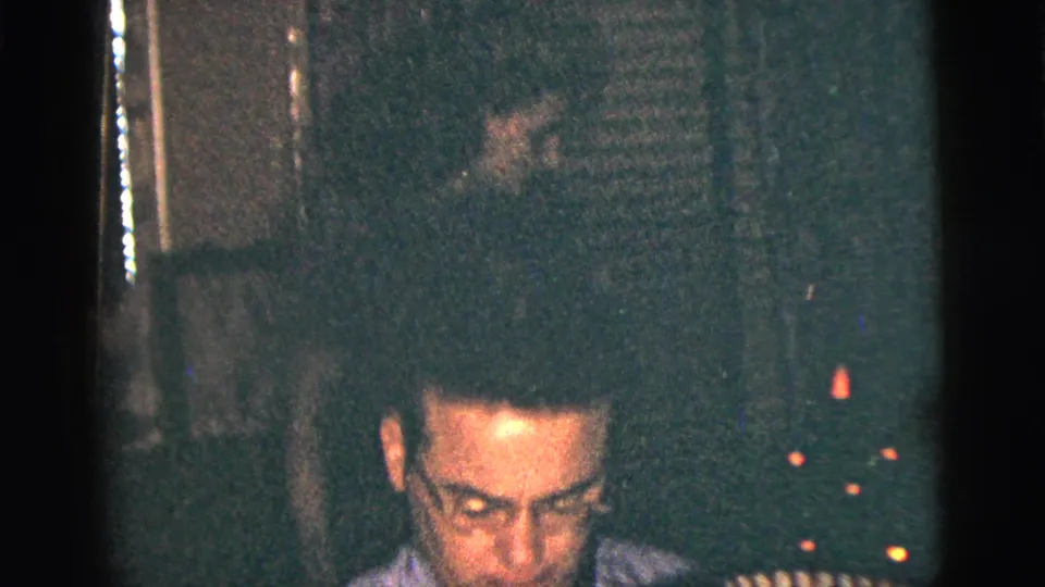 The camera catches the accordion player low in frame, with shadowed background detail and warm points of light in the private room.