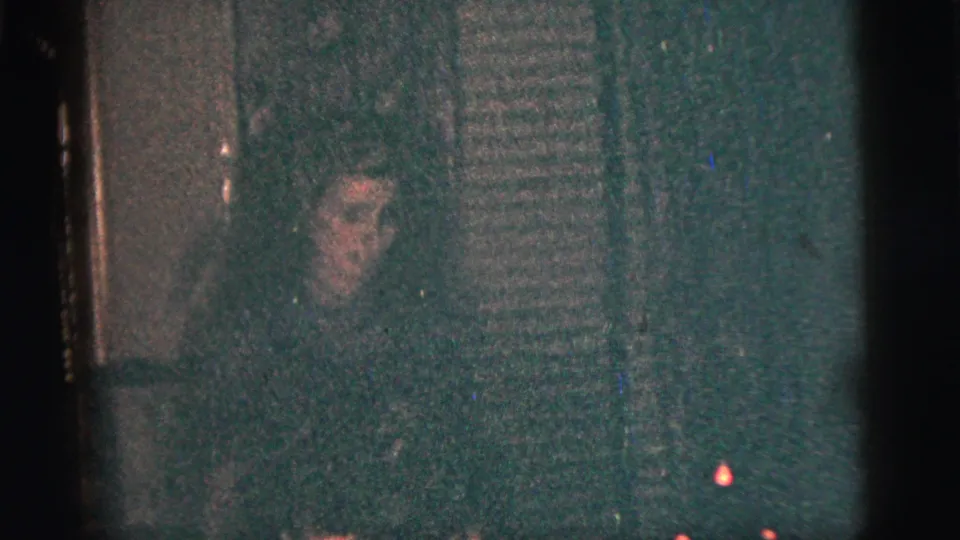 A dim 1956 New York City home-movie frame shows a person watching near vertical interior texture, with the accordion player mostly out of view.