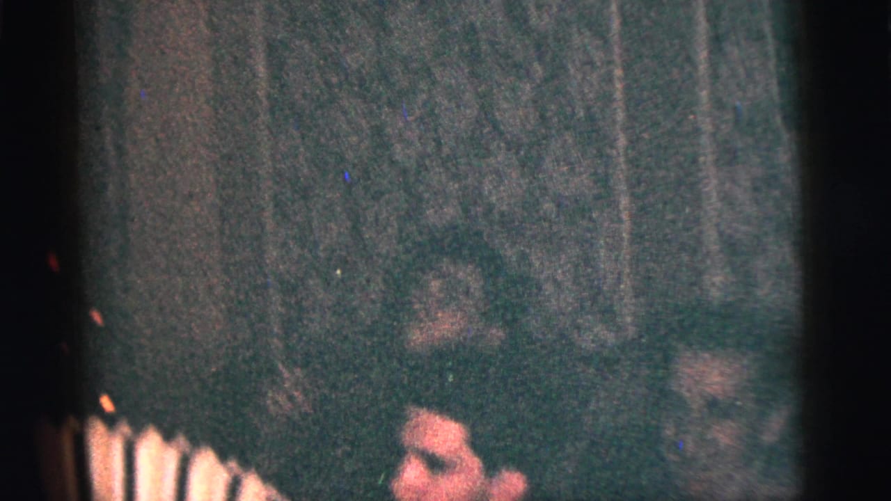 Young man seated indoors playing a small accordion while other men sit nearby.