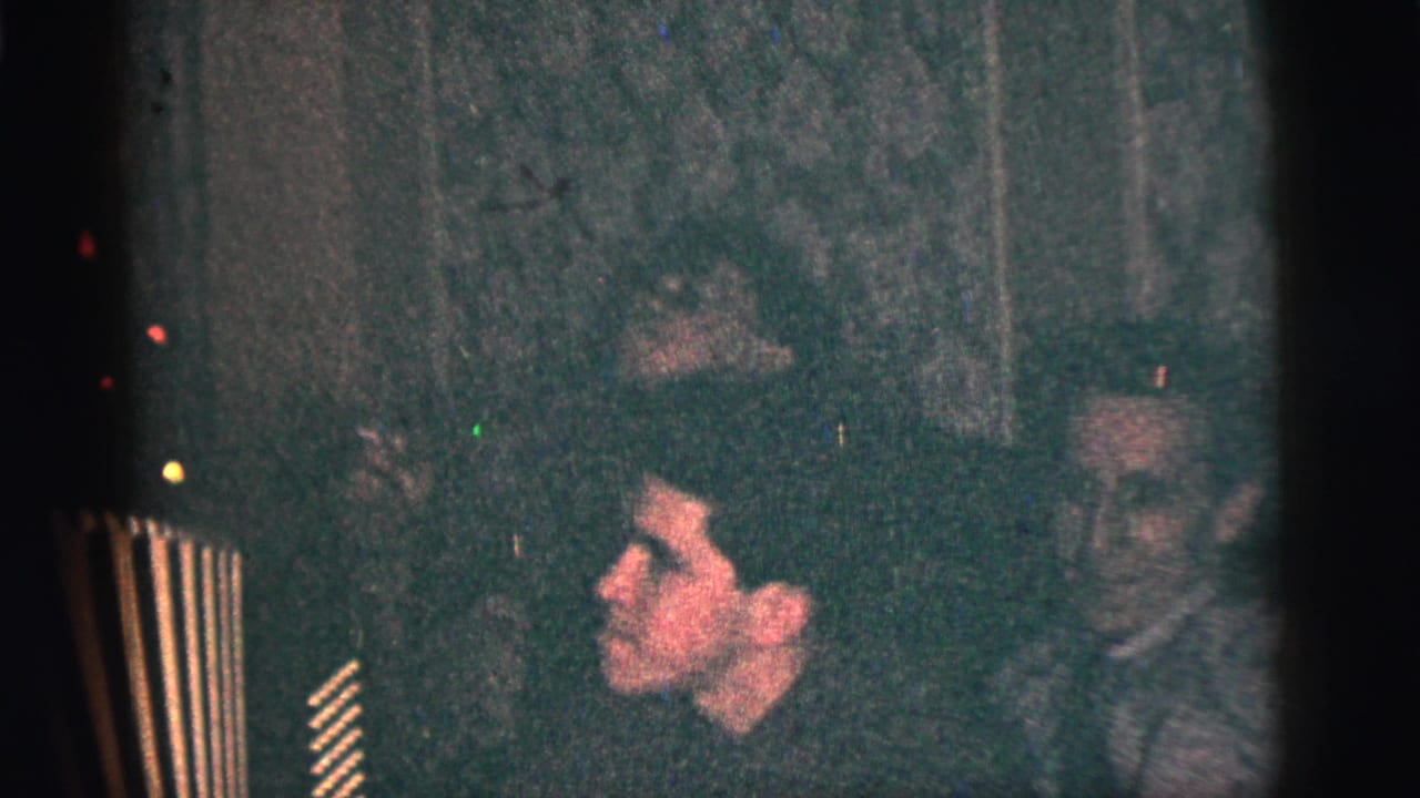 1956 New York City frame showing men seated close together in a dim room, with one face in profile and another man watching nearby.