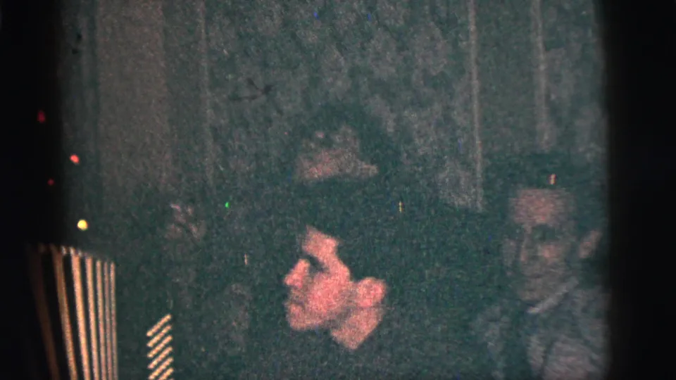 1956 New York City frame showing men seated close together in a dim room, with one face in profile and another man watching nearby.