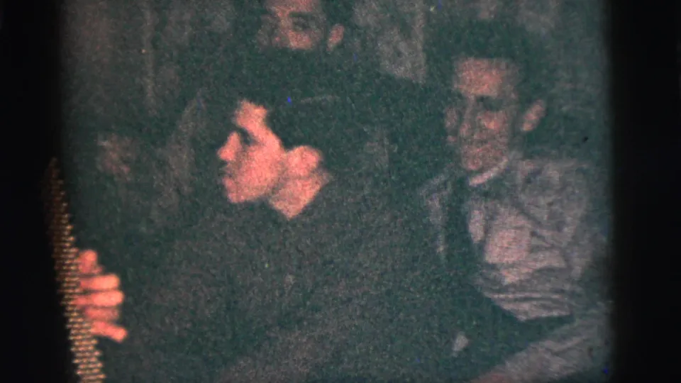 1956 New York City home-movie frame with an accordion player at left and another seated man watching in a dark private interior.