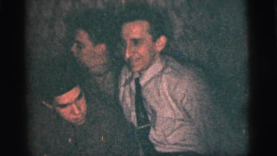 1956 New York City frame of several men seated closely in a dim private room, including a smiling man in a light shirt and tie.