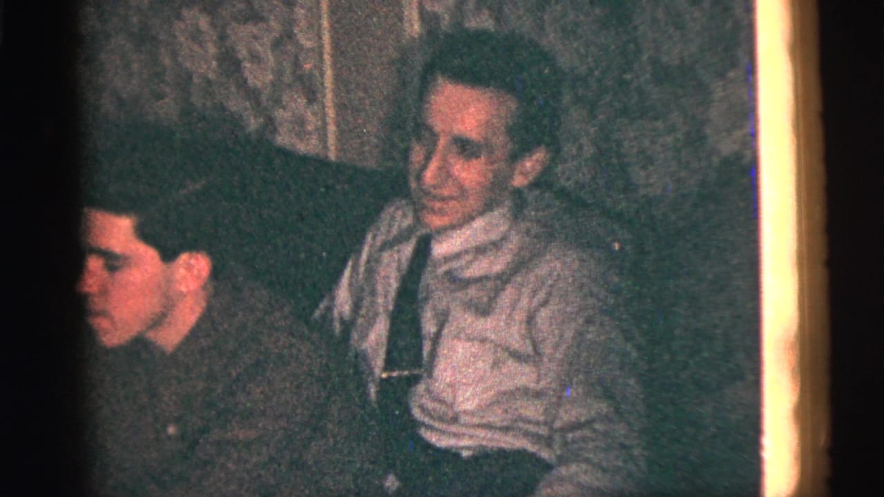 1956 New York City home-movie frame showing seated men watching from a dim indoor gathering, with soft 8mm color and grain.