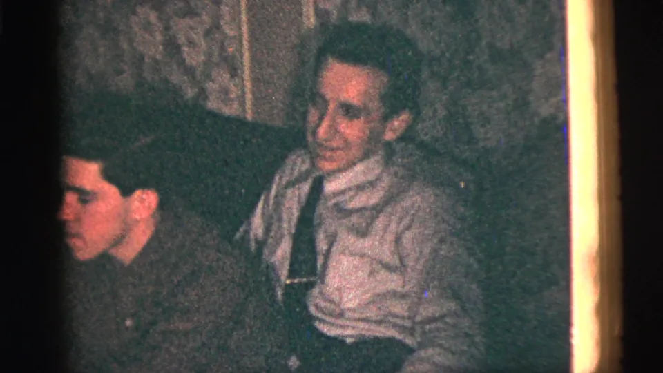 1956 New York City home-movie frame showing seated men watching from a dim indoor gathering, with soft 8mm color and grain.