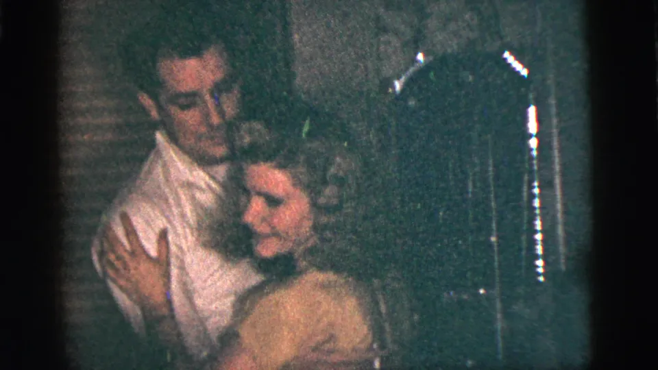1956 New York City home-movie frame of the couple continuing a close dance in a dim interior with visible film grain.