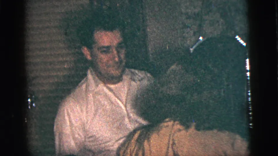 A young man in a pale shirt looks toward the camera while his dance partner in a yellow dress moves across the foreground.