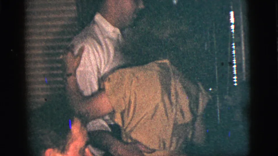 A man in a white collared shirt holds a woman as they dance in a dim 1956 New York City home interior.