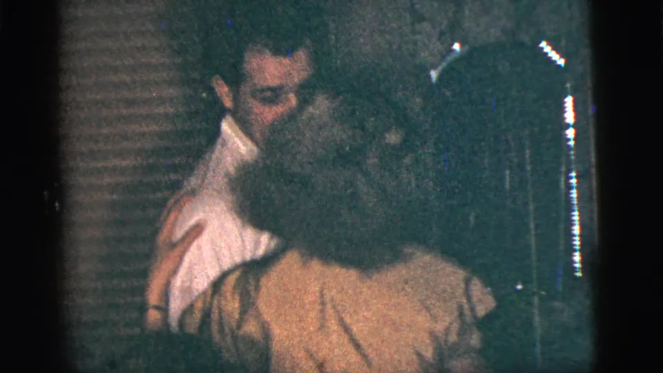 The couple remains in a close dance hold inside a dark New York City home interior, with blinds or paneling behind them.
