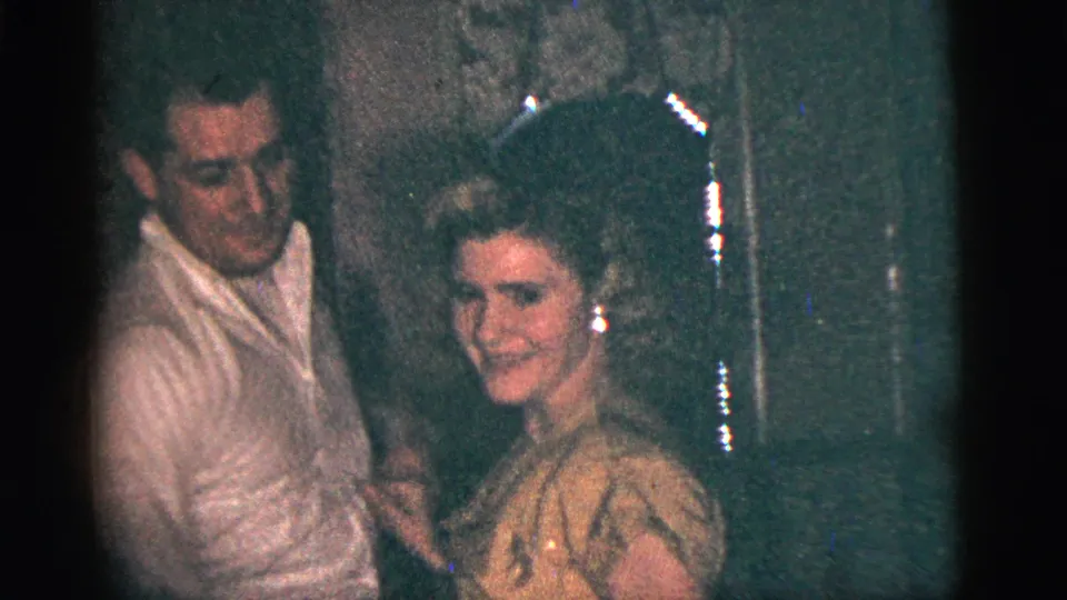 A smiling woman in a light dress faces the camera while dancing with a man in 1956 New York City home-movie footage.
