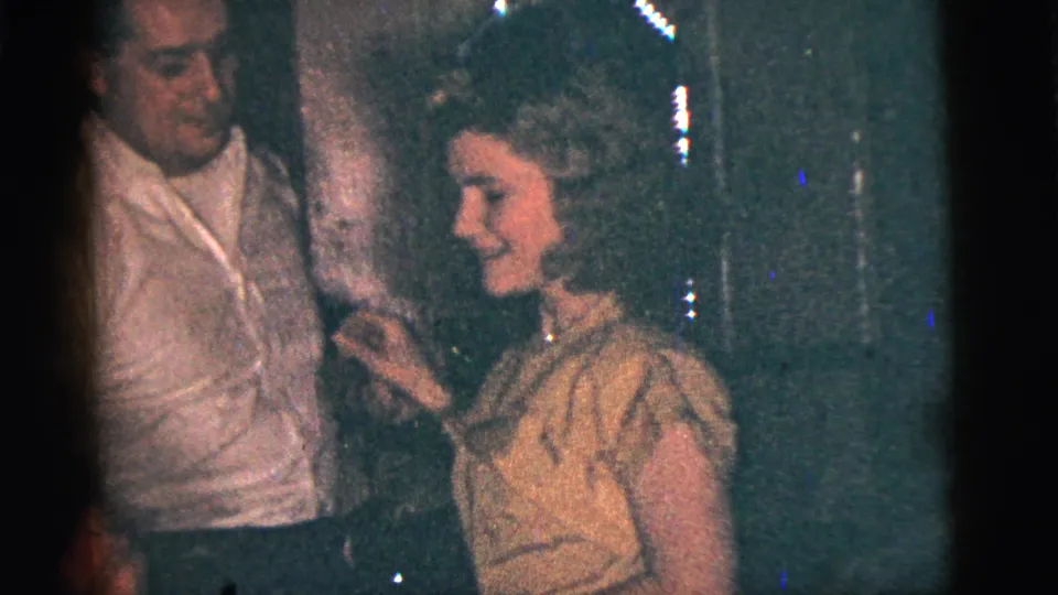 In 1956 New York City home-movie footage, a man in a white shirt and a woman in a light dress dance in a dark indoor gathering.