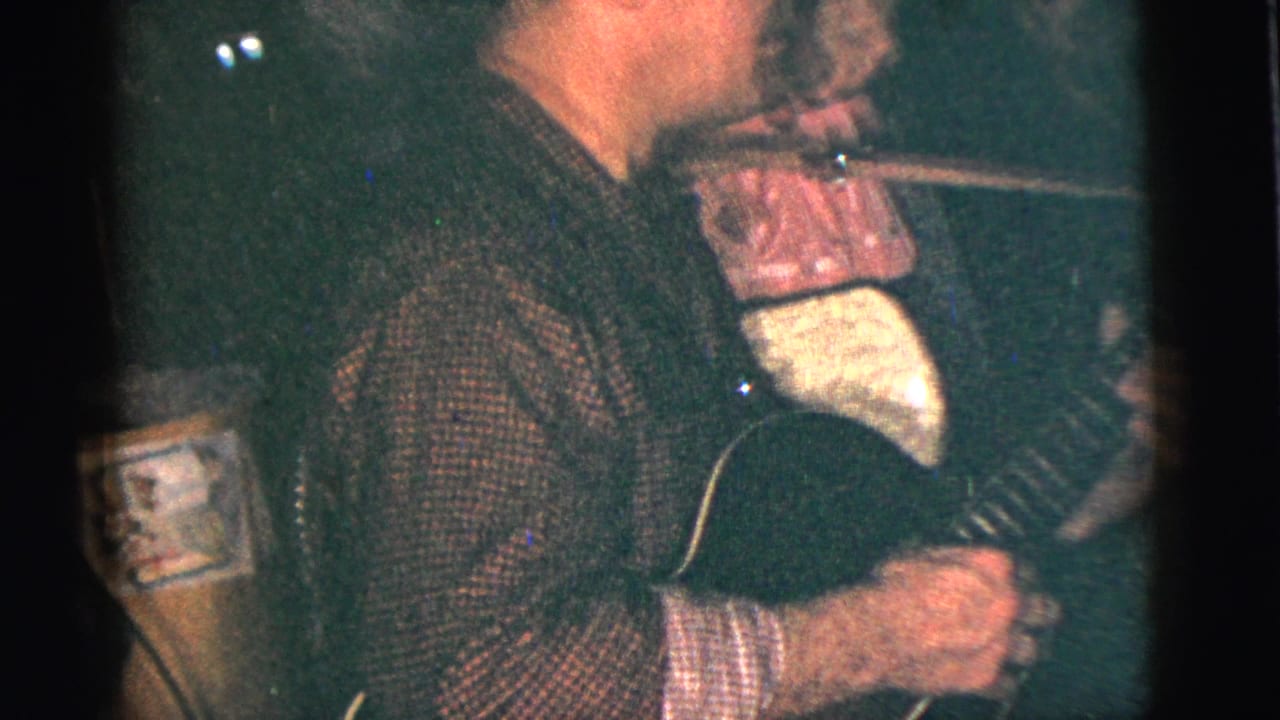 A close, grainy 8mm view shows a guitarist's torso and hands with the instrument during the indoor social scene.