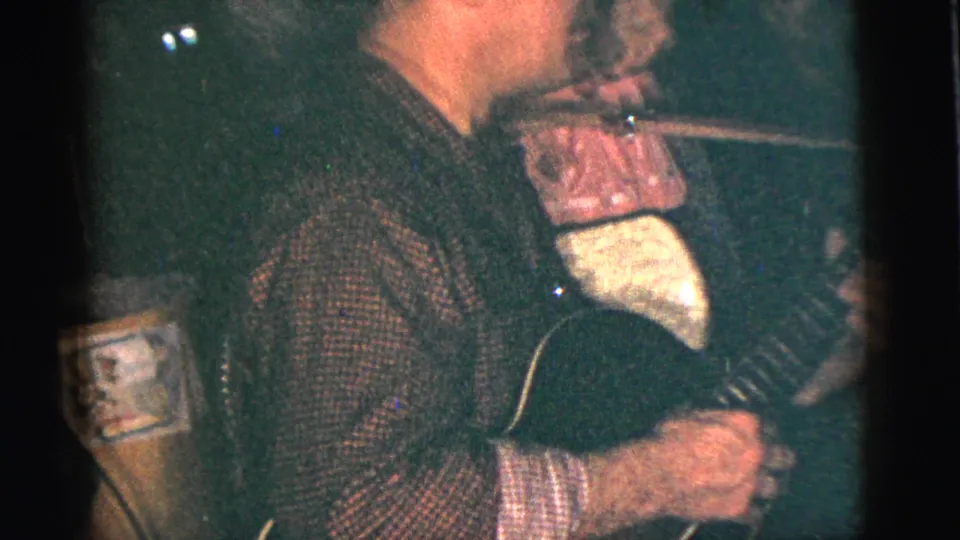 A close, grainy 8mm view shows a guitarist's torso and hands with the instrument during the indoor social scene.