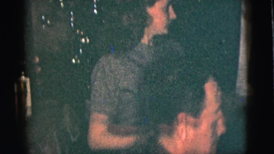 A woman dances in profile inside the dim 1956 New York City home-movie scene, with another adult partly visible nearby.