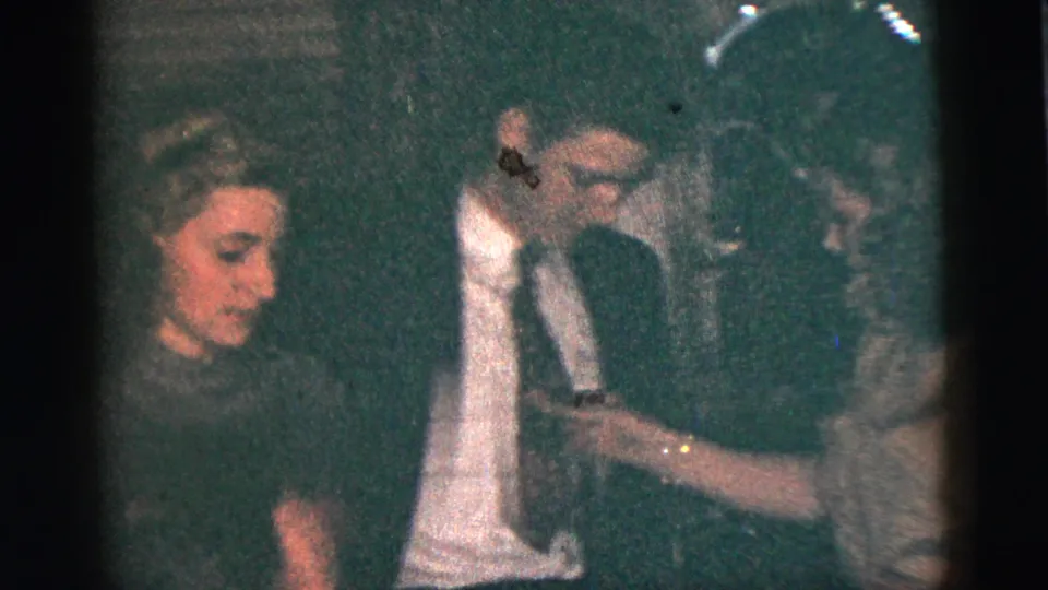 Several adults appear together in the low-light 1956 New York City gathering, including a seated woman, a standing man, and another person at right.