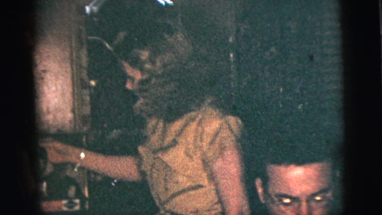 Woman in a dress stands near seated party guests in a dim room.