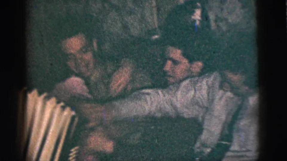 Two adult men appear in close conversation or song near an accordion during the metadata-supported 1956 New York City party footage.