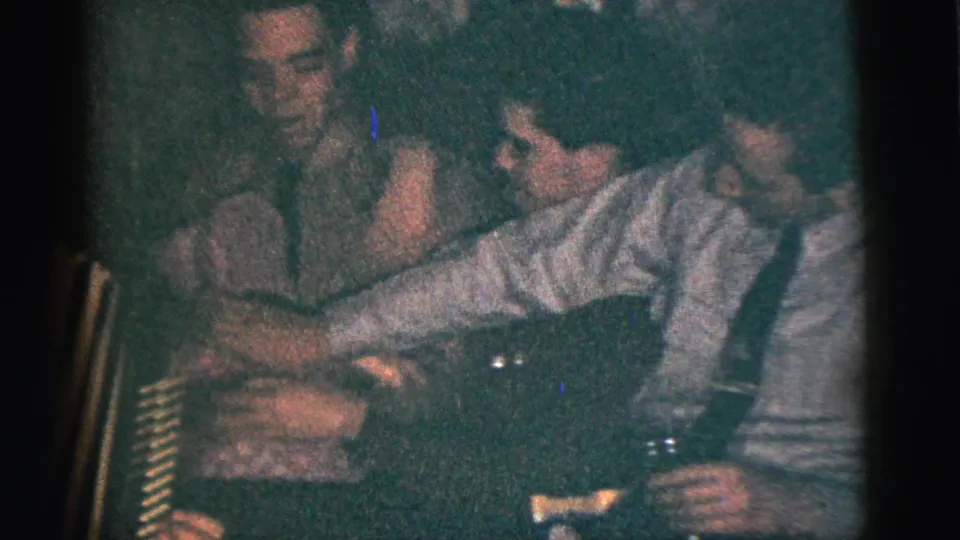 Adults sit close together at a dim 1950s New York City gathering while one guest reaches across toward someone near the accordion.