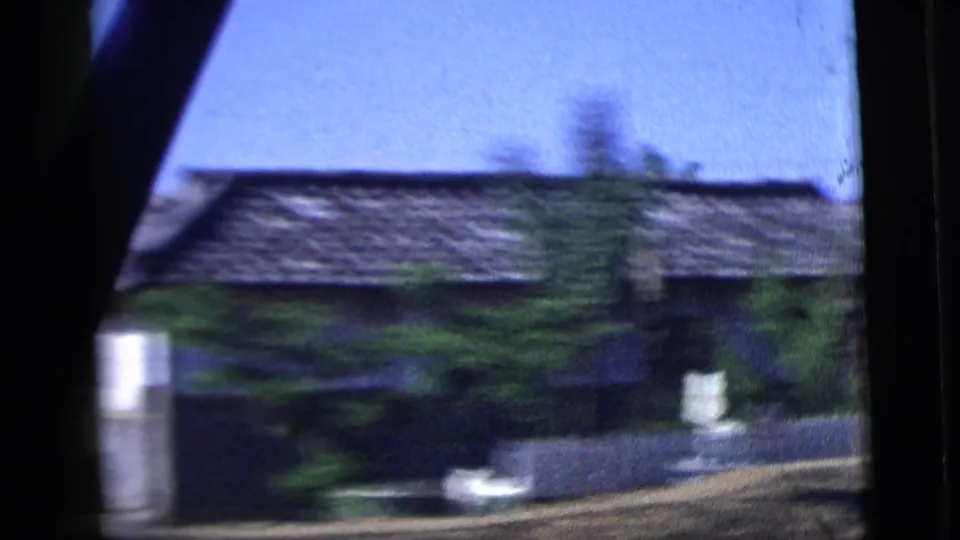 1958 8mm car-window view in the New York City metadata context, showing a low house, wall, trees, and motion blur.