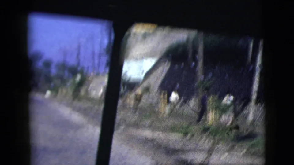 Blurred 8mm frame from inside a moving car, showing a narrow road, white walls, a dark roof, posts, and vegetation.