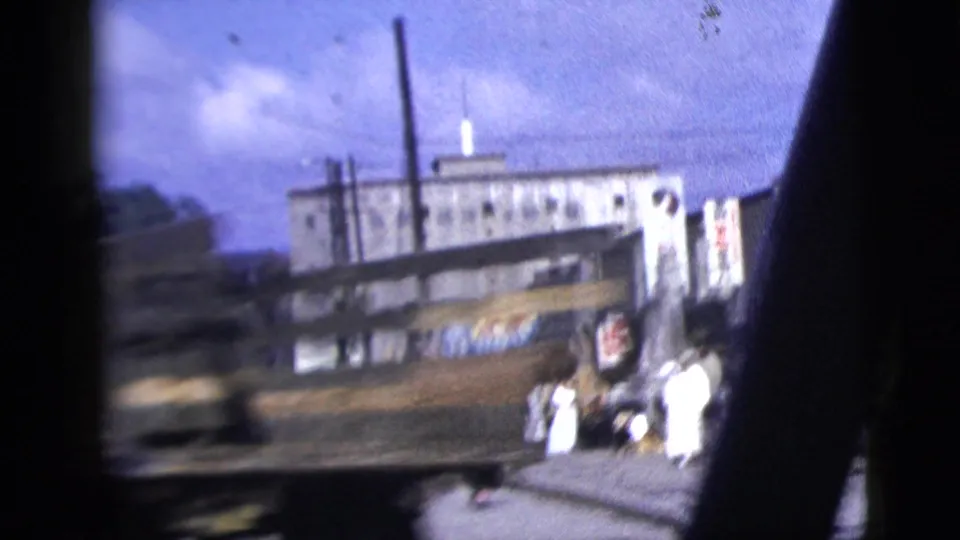 Drive-by frame of a New York City street corner in 1958, with pedestrians near a pale low-rise building and a blurred passing vehicle.