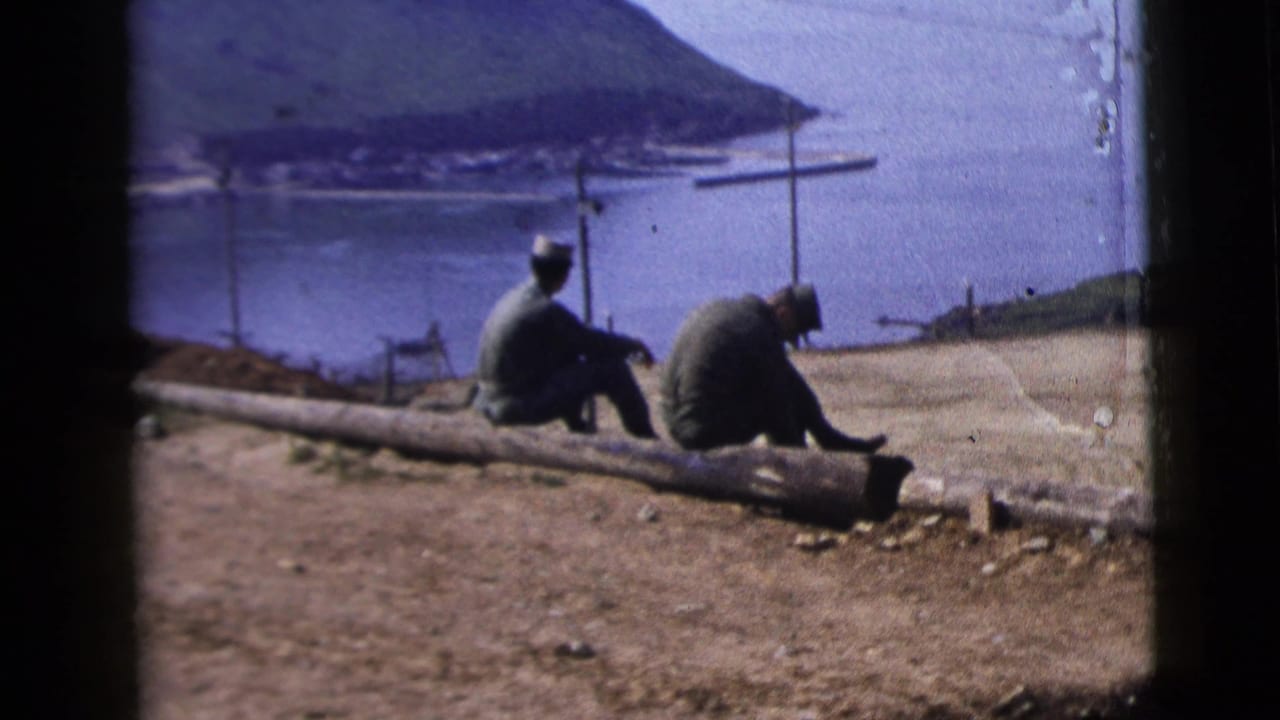 Two adult men in military-style clothing sit on a log at a dirt overlook above a coastal bay, from 1958 8mm home-movie footage.