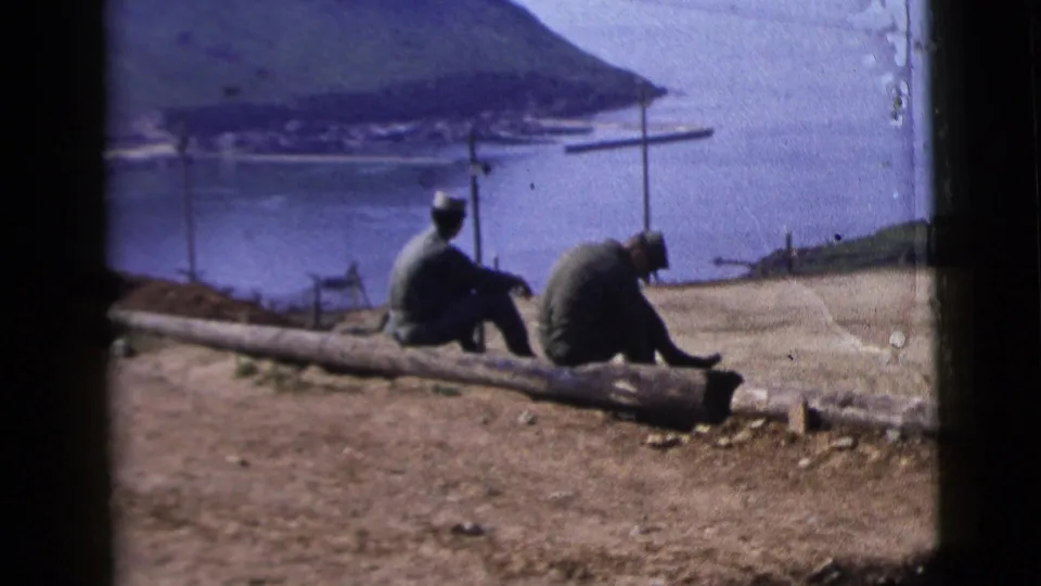 Two adult men in military-style clothing sit on a log at a dirt overlook above a coastal bay, from 1958 8mm home-movie footage.