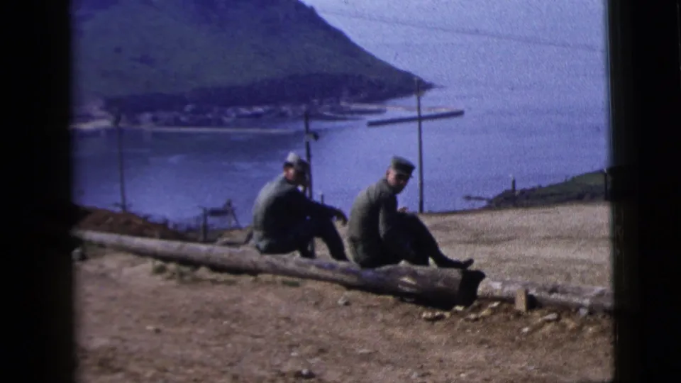 Two adult men on a hillside log turn slightly toward the camera, with bay water, mountains, and shoreline behind them.