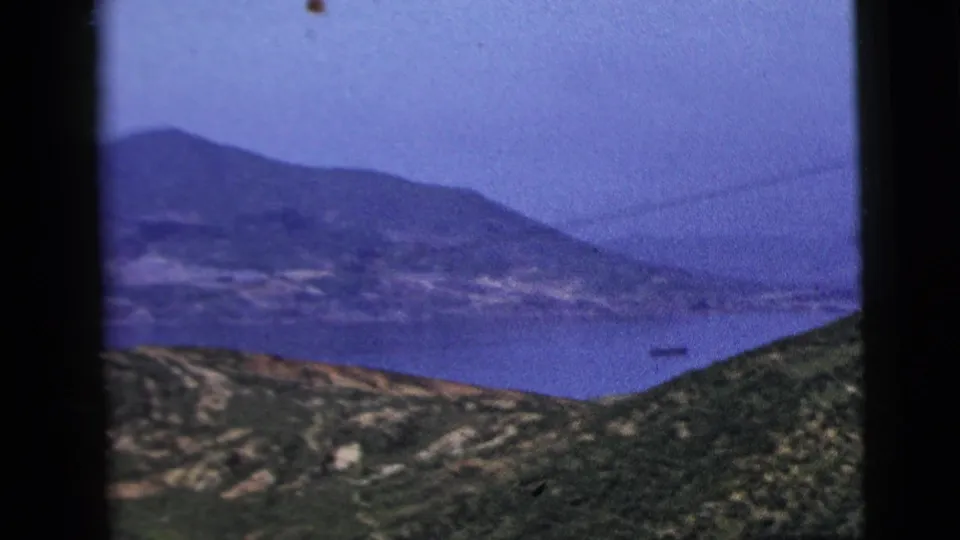 The shot opens to a wider 1958 8mm view of a coastal bay, green slopes, mountains, and a small distant vessel or structure on the water.