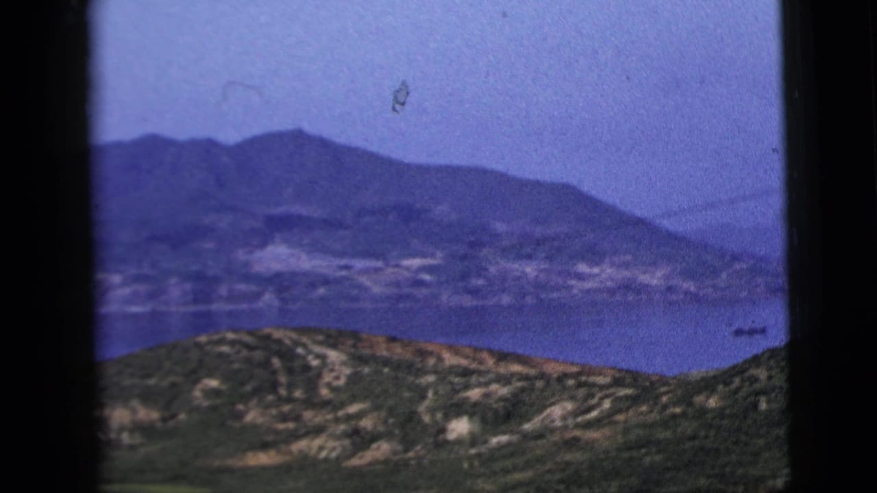 Window-framed view of hills, water, distant mountains, and small boats under a hazy sky.