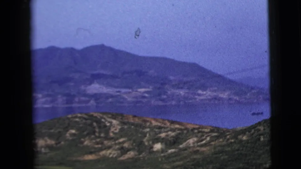 1958 8mm window-framed travel view of rolling hills, a broad water area, distant mountains, and small boats in hazy light.