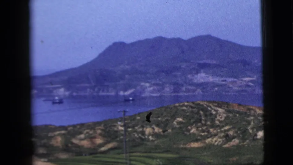1958 8mm view across a broad water area toward mountains, with small boats, shoreline hills, and a foreground utility pole.