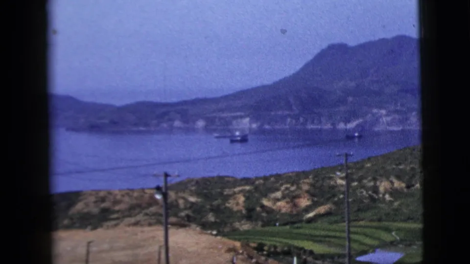 1958 8mm shoreline view with water, distant mountains, small boats, foreground hills, utility poles, and overhead wires.
